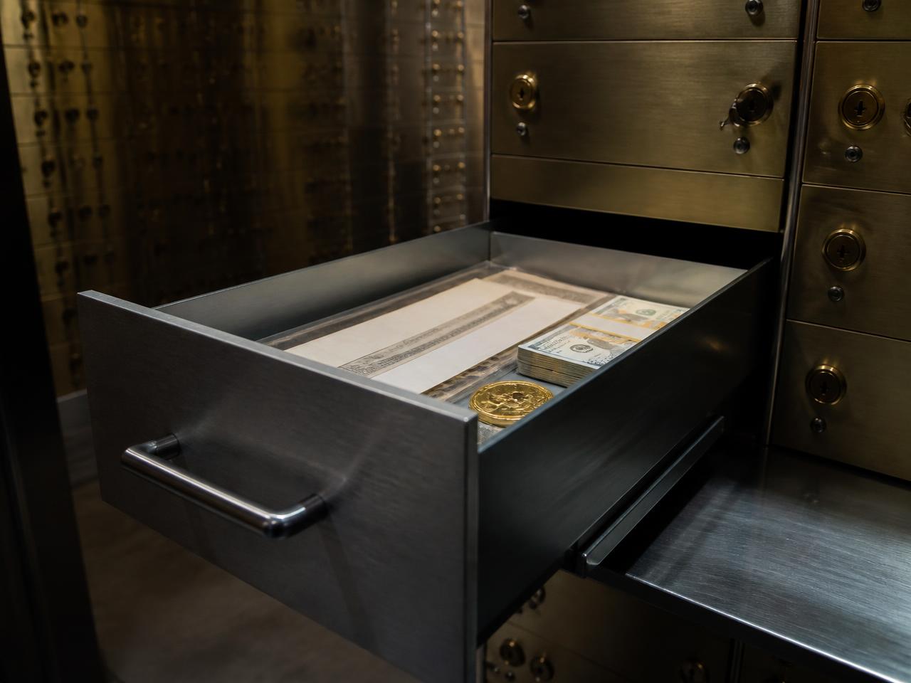 An open safe deposit box holding cash, documents and a gold coin inside a bank vault